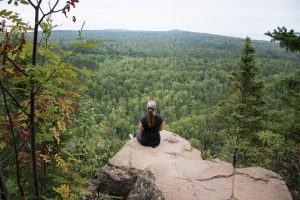 Mountaintop view of the Superior National Forest. There is a woman sitting on a rock on top of a mountain. She is looking at a remote, green forest from her high-up position.
